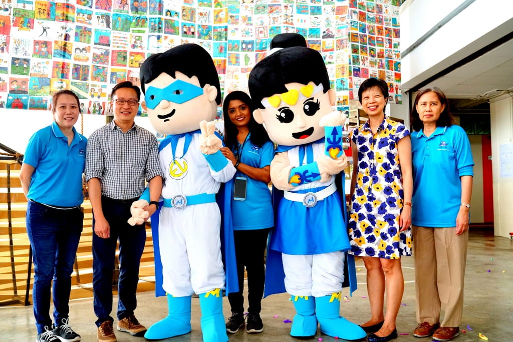 Five people pose with two superhero mascots in blue and white costumes before a colorful wall of children's artwork.